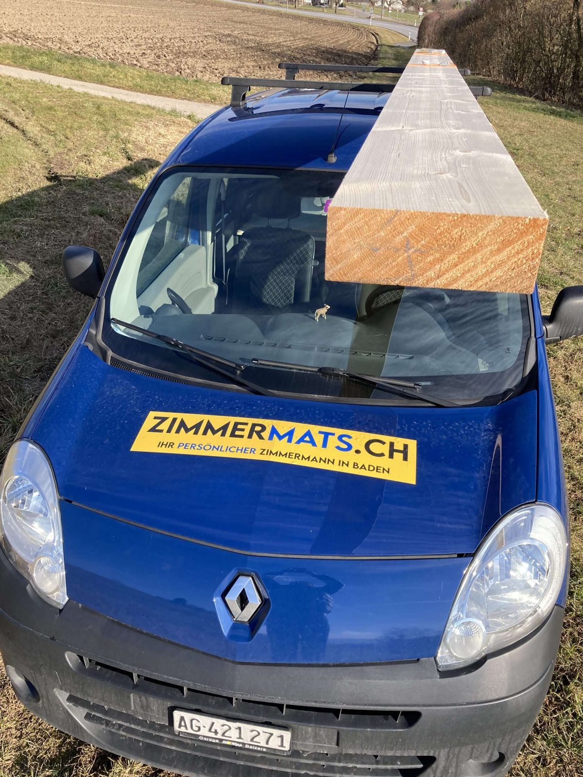 Blauer Firmenwagen mit gelbem Schild ZIMMERMATS.CH und Balken auf dem Dach